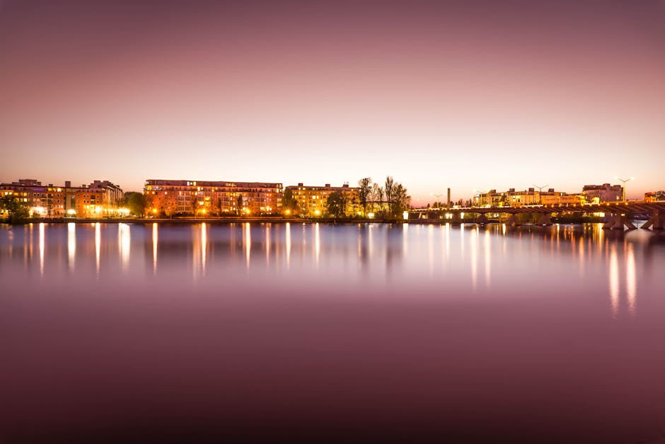 A blurred city skyline at dusk with glowing lights and calm water.